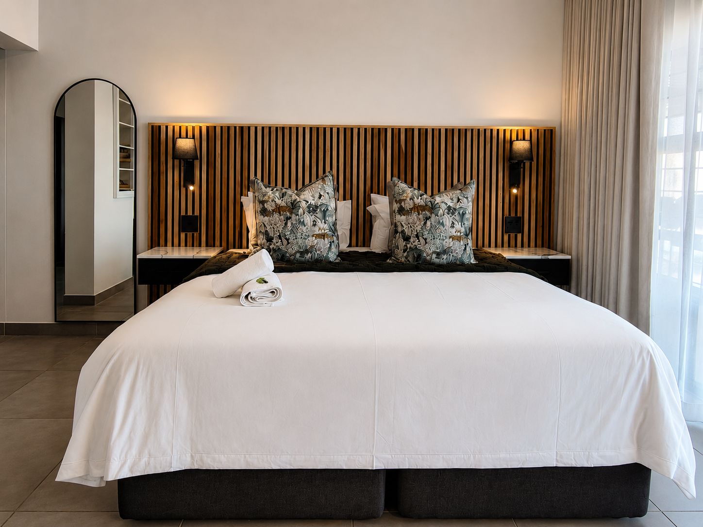 A modern hotel room with a large bed, two bedside tables, a mirror, and a wooden panel wall.