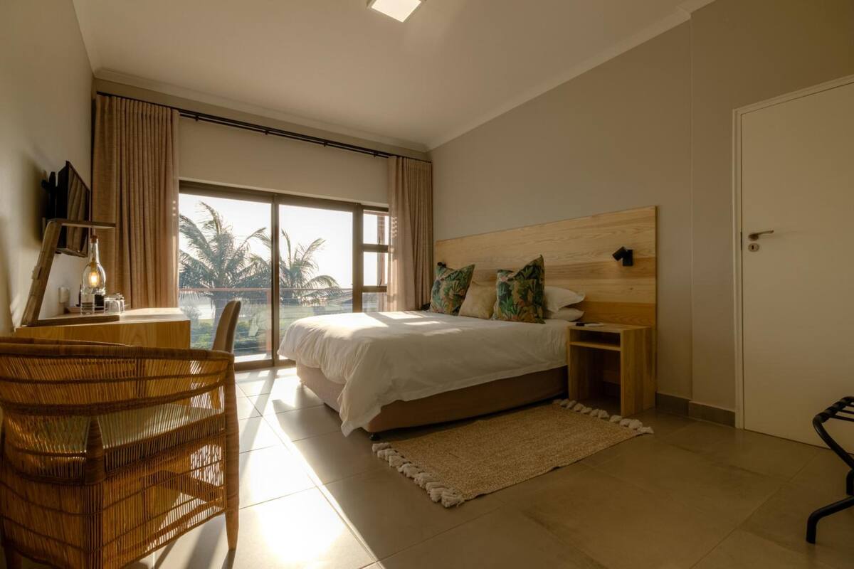 A bedroom with a large bed, a desk, a chair, and a view of palm trees through a sliding door.