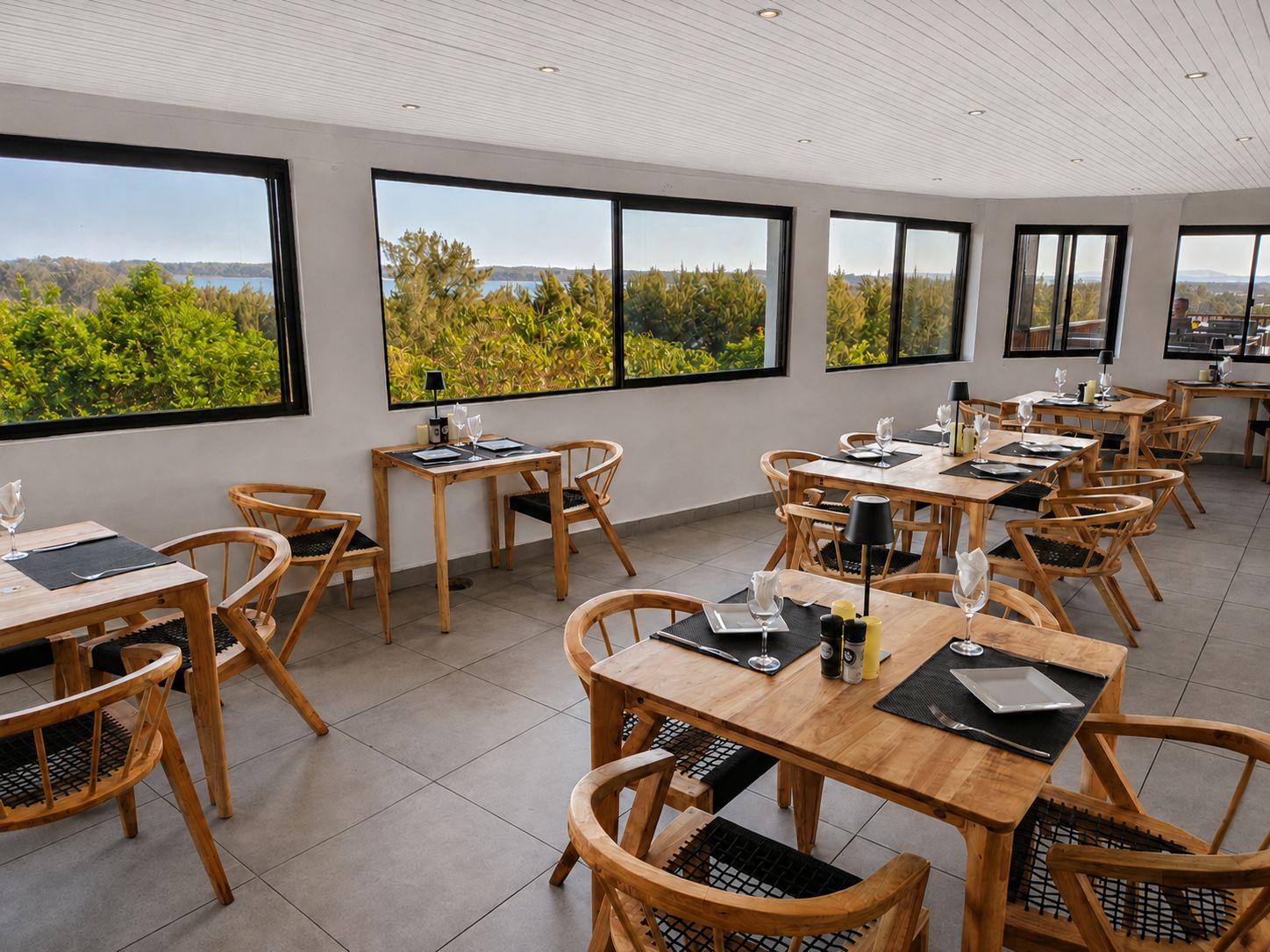 A dining area with multiple tables and chairs, large windows with a view of greenery and hills, and a ceiling with recessed lighting.