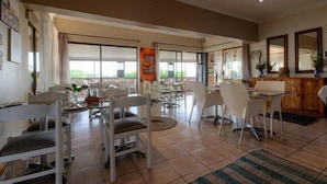 A spacious dining area with multiple tables and chairs, large windows providing natural light, and a view of the outdoors. - The Ridge Guesthouse (Richards Bay)