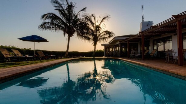 A swimming pool with lounge chairs and umbrellas, surrounded by palm trees and a building with outdoor seating. - The Ridge Guesthouse (Richards Bay)