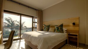 A hotel room with a large bed, a wooden headboard, a nightstand, a chair, and a view of palm trees through a sliding glass door. - The Ridge Guesthouse (Richards Bay)