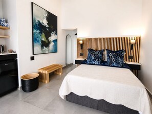A modern bedroom with a large bed, a wooden headboard, bedside lamps, a bench, a trash bin, and a wall-mounted artwork. - The Ridge Guesthouse (Richards Bay)