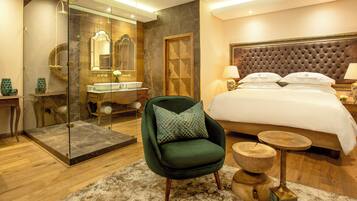 Una habitación de hotel moderna con una cama grande, un sillón verde, una mesita de madera, un área de ducha con mampara de vidrio y un piso con mosaicos de mármol.