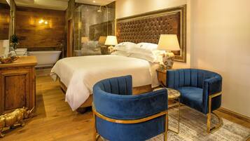 Quarto de hotel com uma cama grande, duas poltronas azuis, cômoda de madeira, banheira e chuveiro com box de vidro.
