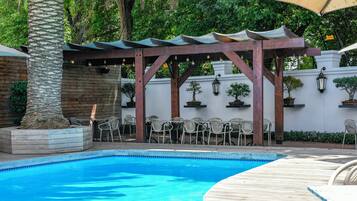 Un espace piscine doté d’une terrasse en bois, de chaises et de tables, le tout abrité par une pergola.