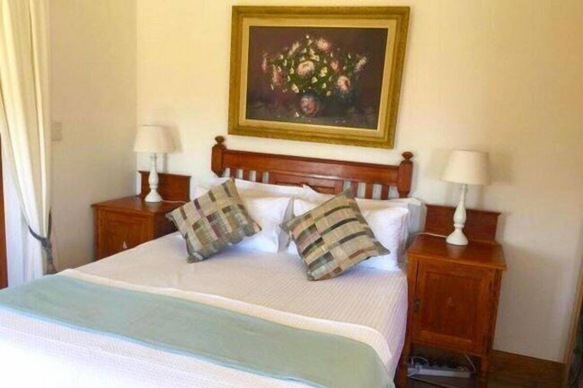 A neatly made bed with a white comforter and two pillows, flanked by wooden nightstands with lamps. A framed painting of flowers hangs above the bed.