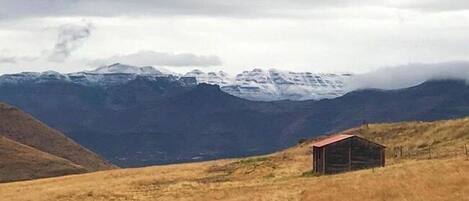 A small wooden cabin in a grassy field with a mountain range in the background.