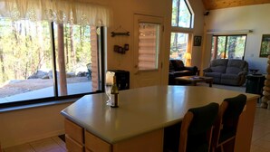 Eine gemütliche Küche mit einer großen Kochinsel, ein Essbereich mit einem karierten Sofa und Blick auf ein bewaldetes Gebiet durch die Fenster.