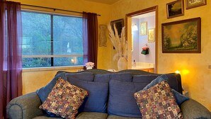 A cozy living room with a large blue sofa, patterned cushions, a window with curtains, and framed artwork on the walls.