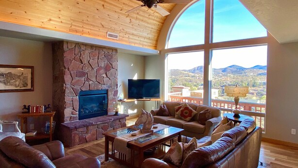 A spacious living room with a stone fireplace, leather furniture, a television, and a large window offering a mountain view. - Longview Lodge - Prescott Cabin Rentals (Prescott)