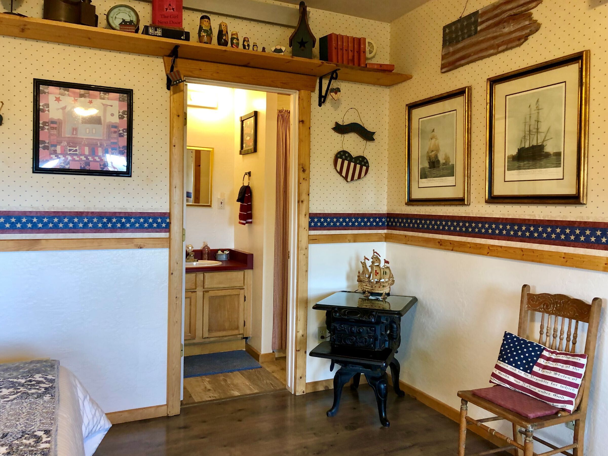 A room with a wooden floor, a small table with a ship model, a chair with an American flag pillow, and a wall with framed pictures and a heart-shaped decoration.