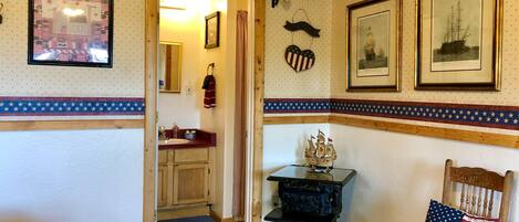 A room with a wooden floor, a small table with a ship model, a chair with an American flag pillow, and a wall with framed pictures and a heart-shaped decoration.