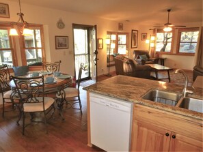 A kitchen with a dishwasher, wooden cabinets, a granite countertop, a dining area with a round table and chairs, and a view of the outdoors through large windows. - Western Pines Cottage - Prescott Cabin Rentals (Prescott)