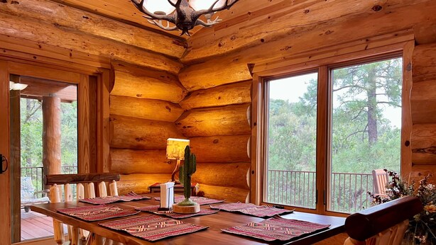 A wooden cabin interior with a dining table, chairs, a cactus centerpiece, and large windows offering a view of the forest. - Timbertrail Lodge - Prescott Cabin Rentals (Prescott)