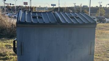 Un contenedor metálico grande y rectangular con techo corrugado, ubicado sobre una superficie de grava en un espacio abierto con césped escaso y cielo despejado.