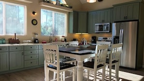 Een keuken met lichtgroene kastjes, een witte eettafel met vier stoelen, een roestvrijstalen koelkast en een magnetron boven het fornuis.