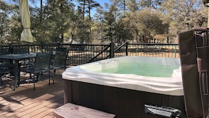 Een hottub op een houten terras met een tafel en stoelen, omringd door een hek en bomen.