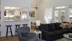 Ein geräumiges Wohnzimmer mit einem schwarzen Sofa, einem grauen Sessel und einem kleinen Beistelltisch. Im Hintergrund ist ein Essbereich mit einem weißen Tisch und Stühlen zu sehen.