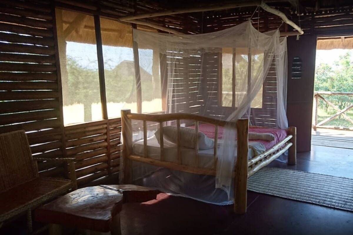 A rustic wooden room with a bed under a mosquito net, a bench, and a view of greenery outside.