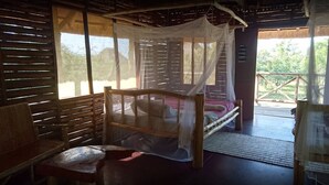 Una habitación rústica de madera con una cama bajo una mosquitera, un banco y vistas al follaje exterior.