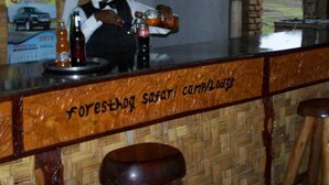 A bar area with a wooden counter and stools. The counter has a sign that reads "Forest Safari Camp Lodge."