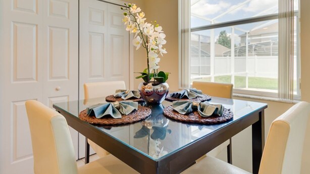 Een eethoek met een glazen tafel, witte stoelen, een vaas met bloemen en uitzicht op een kas door grote ramen.