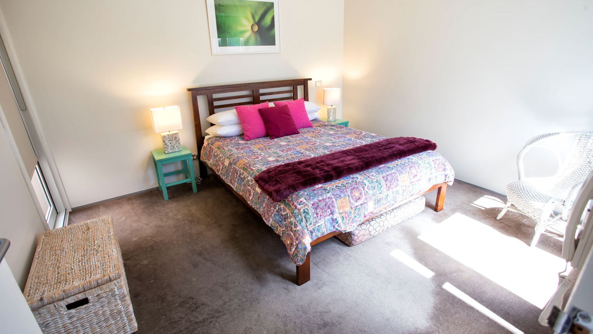 A bedroom with a bed, two bedside tables, a chair, a woven basket, and a framed picture on the wall.