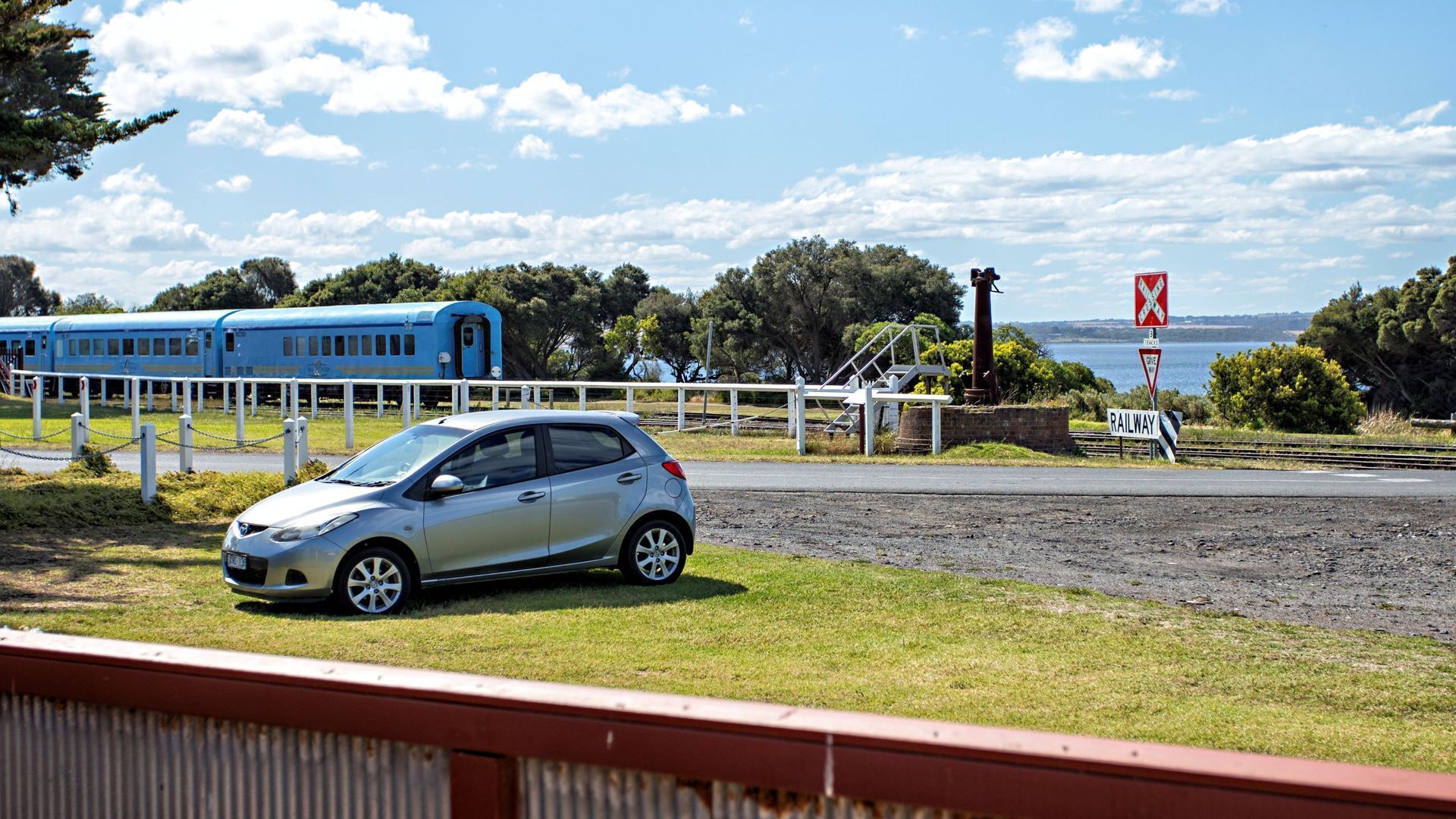 A car parked near a railway crossing with a blue train in the background.