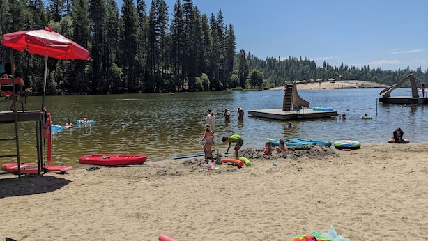 A lakeside beach with a lifeguard chair, a red umbrella, and a sandy area with beach toys and a slide structure in the background. - Dandelion's Lair, a 3 bedroom, 2.5 bathroom cabin in Twain Harte! (Twain Harte)