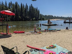 A lakeside beach with a lifeguard chair, a red umbrella, and a sandy area with beach toys and a slide structure in the background.