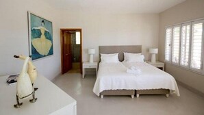 A bedroom with a large bed, bedside tables, a nightstand, a door, and a wall-mounted painting. - Stella Maris (Plettenberg Bay)