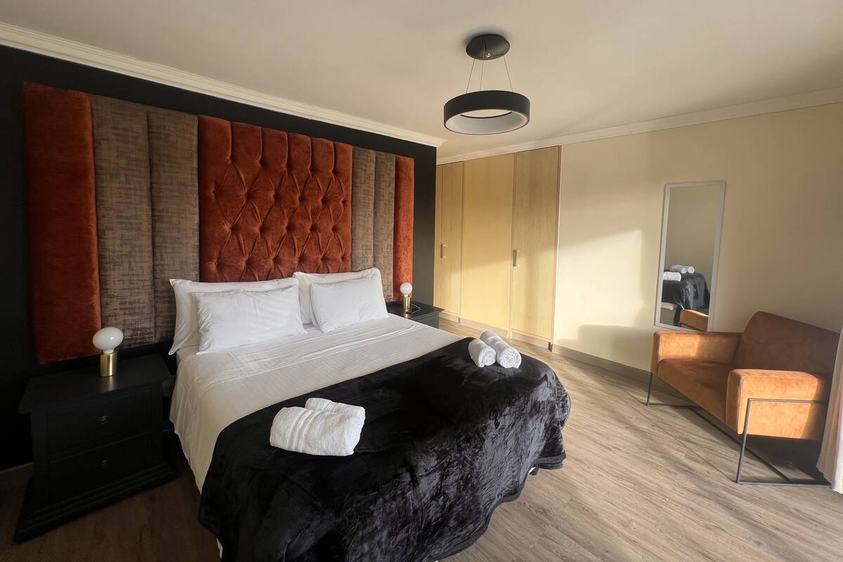 A hotel room with a large bed, a nightstand, a black ottoman, a wooden wardrobe, a mirror, and a seating area with a chair.