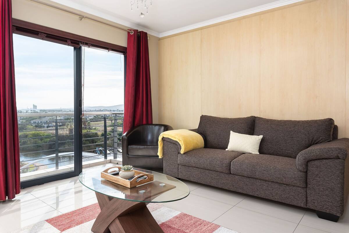 A modern living room with a glass coffee table, a grey sofa, and a view of a cityscape through large windows.