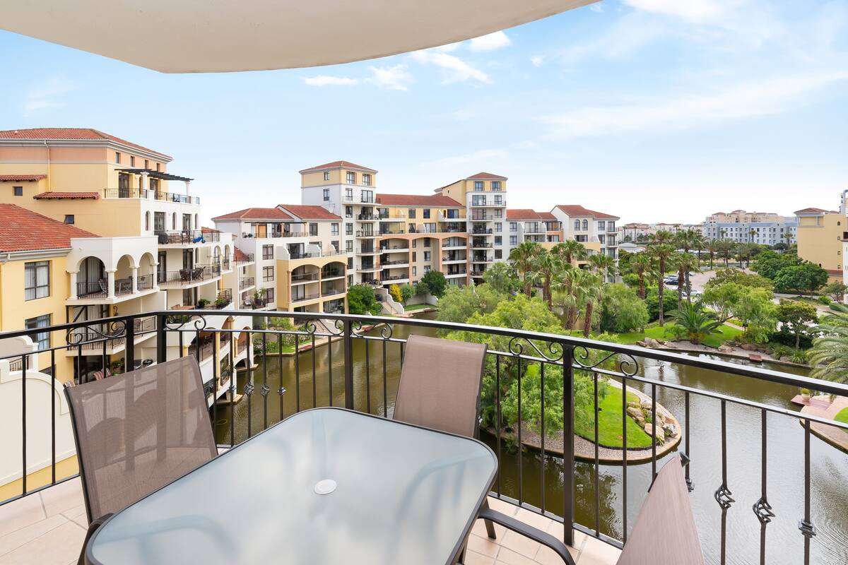 A balcony with a glass table and chairs overlooking a river, surrounded by multi-story buildings with balconies and a garden area.