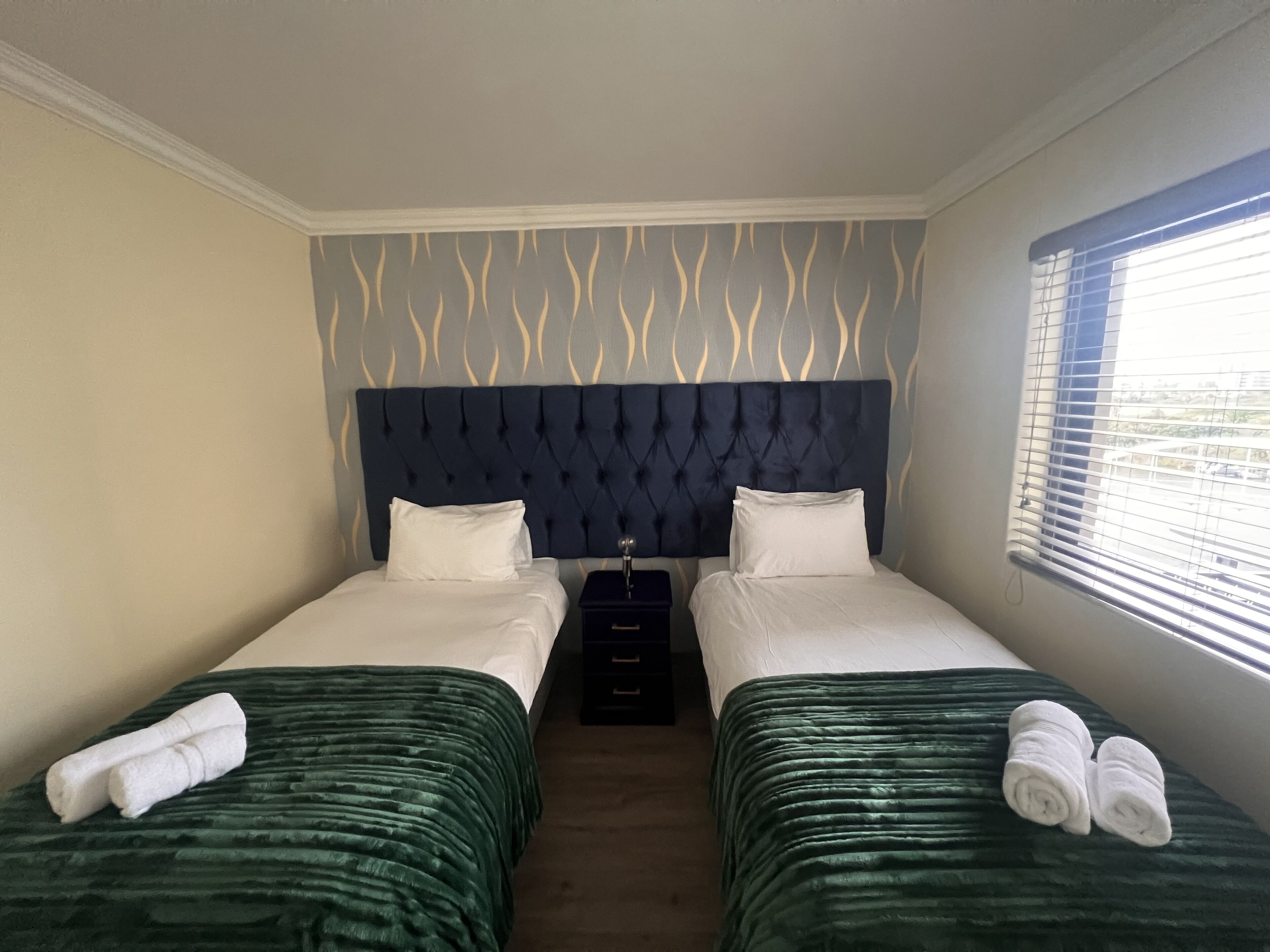 Ein Hotelzimmer mit zwei Betten, einem dunklen Kopfteil, einem Fenster mit Jalousien und einer Wand mit Muster.