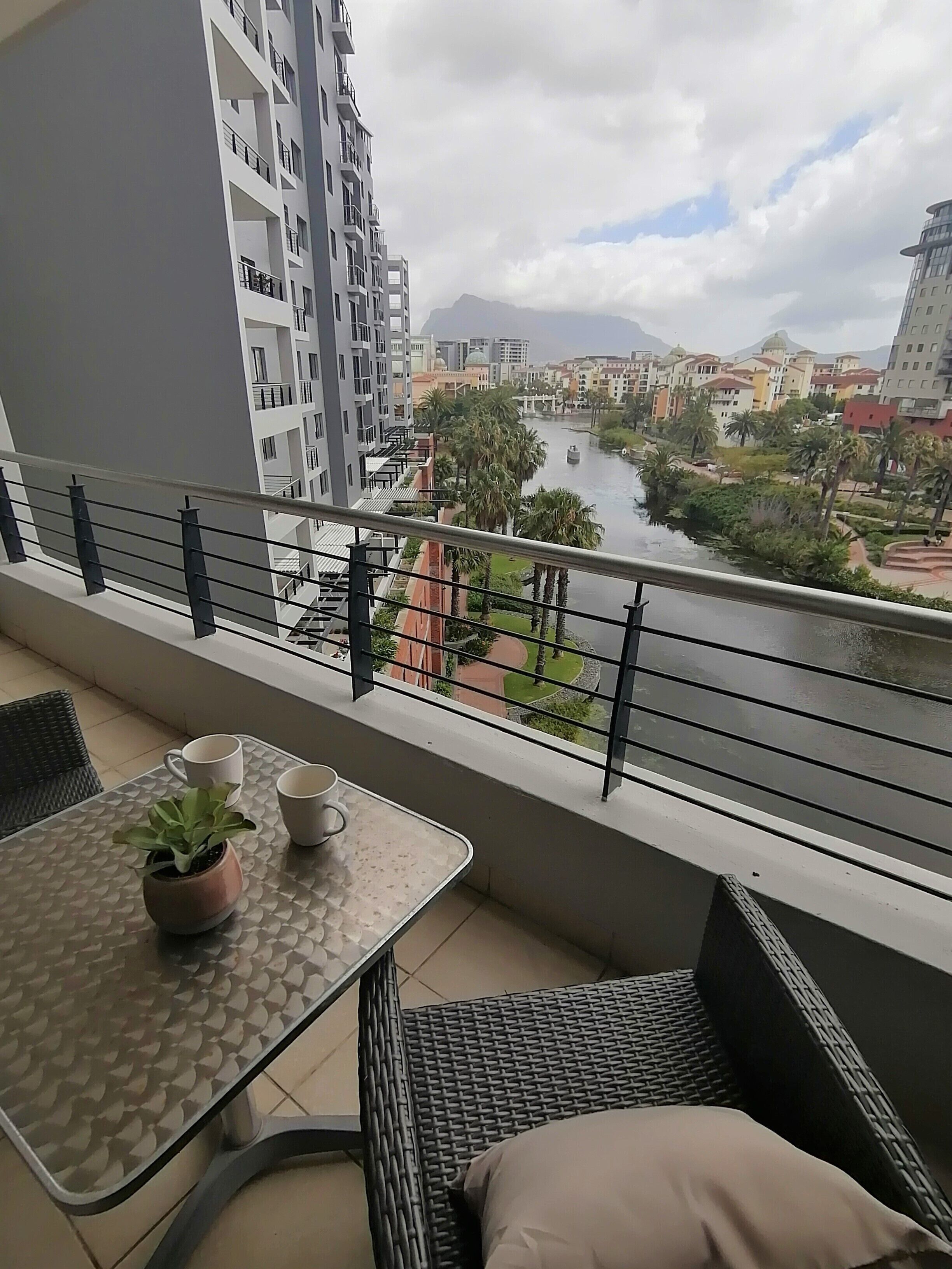Ein Balkon mit Tisch, Stühlen und einem Topfpflanze. Im Hintergrund sind ein Fluss und Gebäude sichtbar.