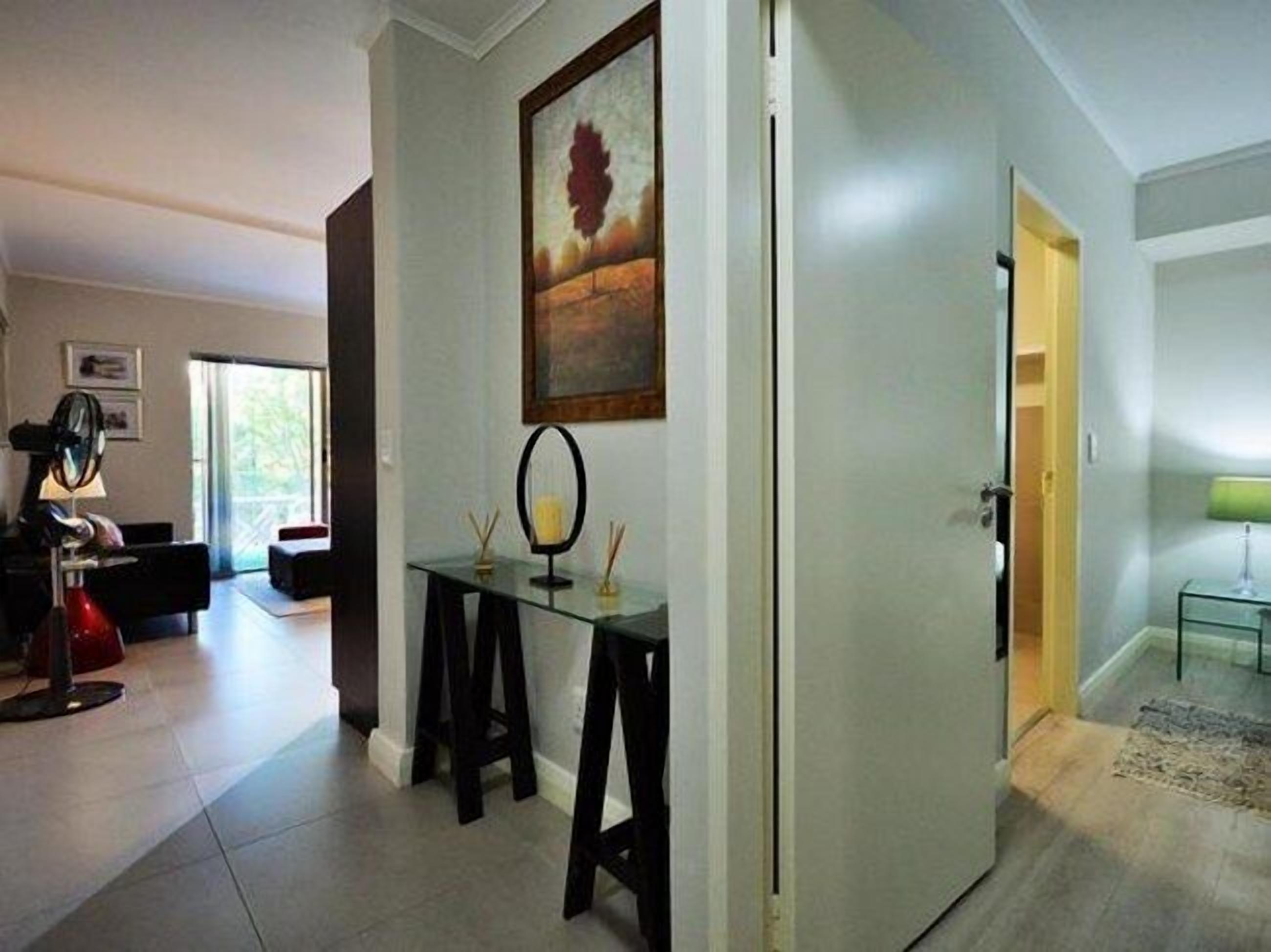 A modern living room with a glass coffee table, a painting on the wall, and a door leading to another room.