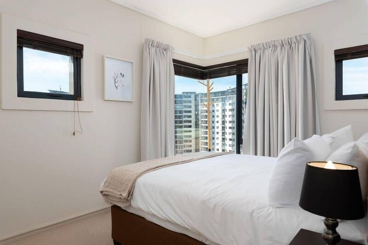 A hotel room with a large bed, a nightstand with a lamp, and a view of high-rise buildings through the windows.