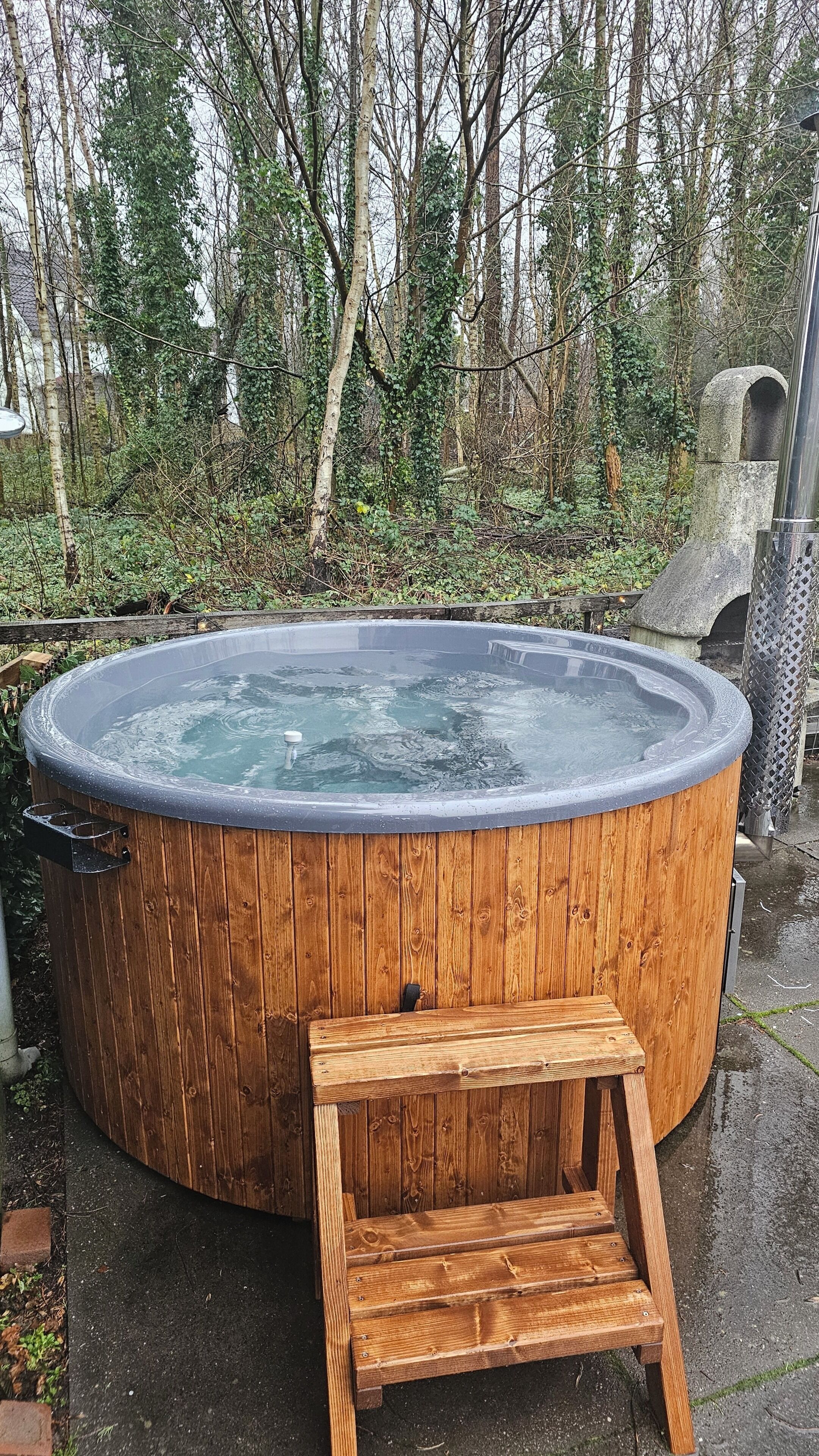 Un bain à remous en bois, avec un banc en bois devant, entouré d’arbres et surmonté d’une structure en pierre en arrière-plan.