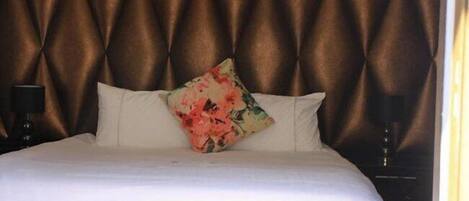 Cama bem arrumada com lençóis brancos e um travesseiro floral, cabeceira marrom escura e duas luminárias de cabeceira.