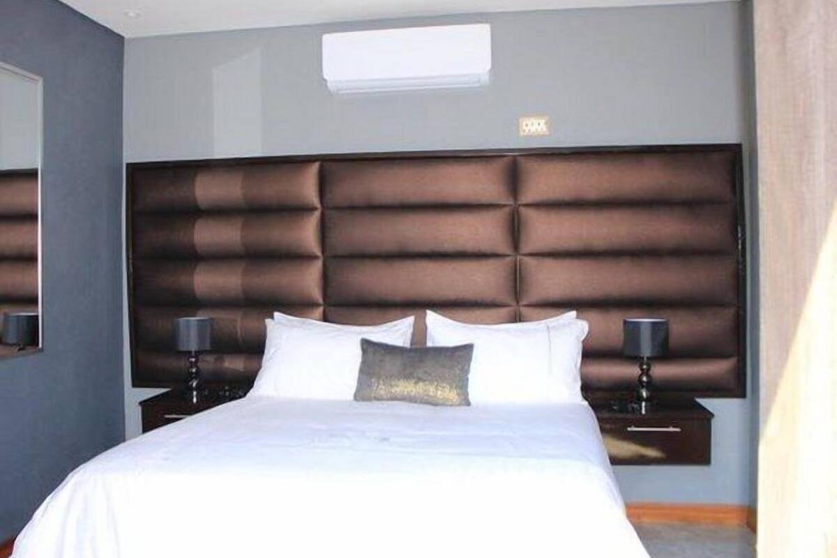 A modern hotel room with a large bed, a padded headboard, two bedside tables with lamps, and a small window above the bed.
