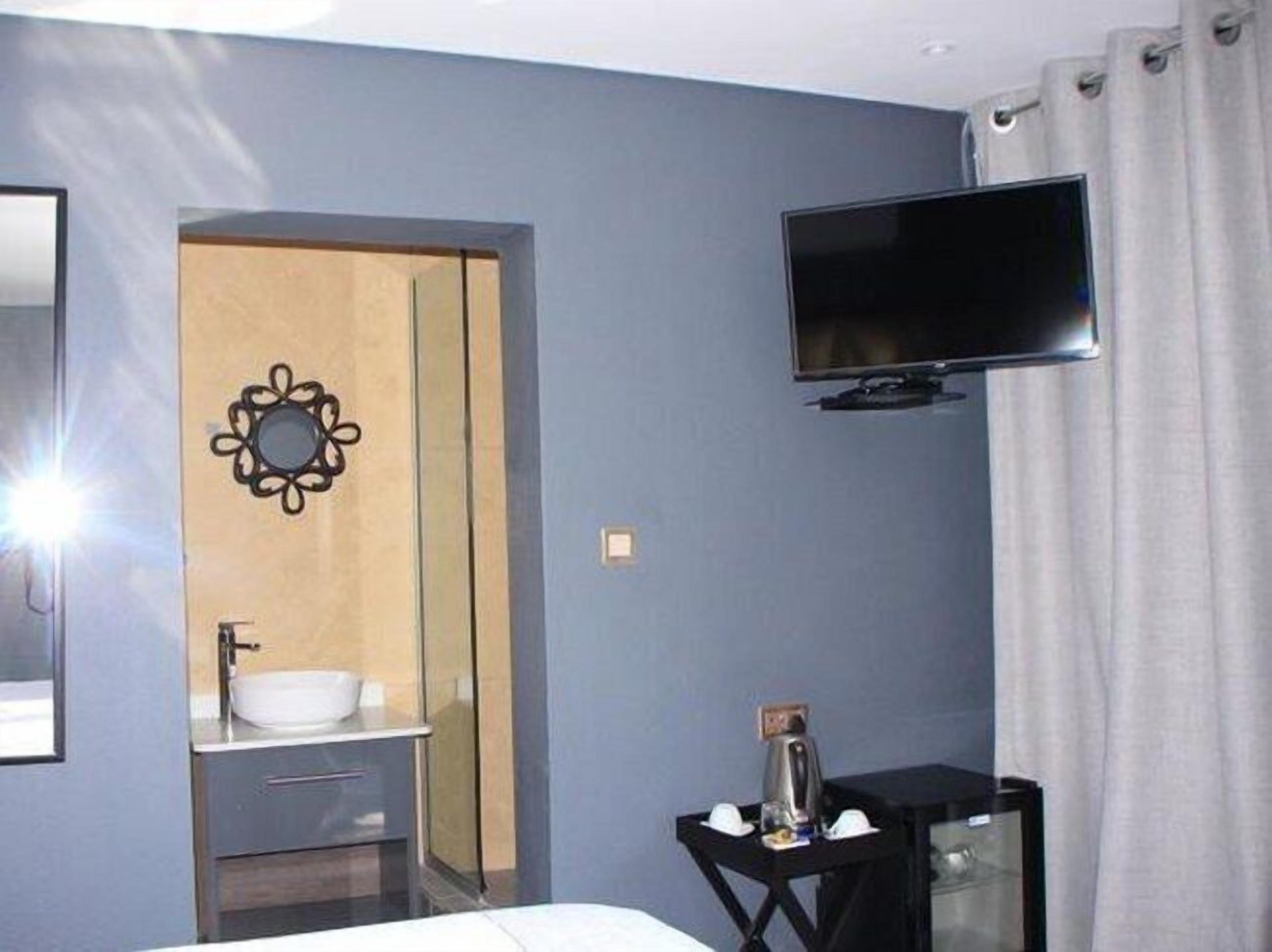 Habitación de hotel con cama, televisor montado en la pared, lavamanos con espejo y una mesita con tetera y tazas.