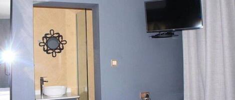 Habitación de hotel con cama, televisor montado en la pared, lavamanos con espejo y una mesita con tetera y tazas.