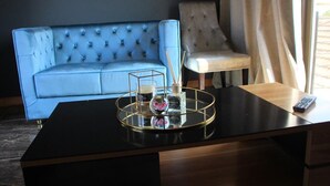 A modern room with a dark blue sofa, a light-colored armchair, and a wooden desk with a tray holding a glass, a bottle, and a candle.
