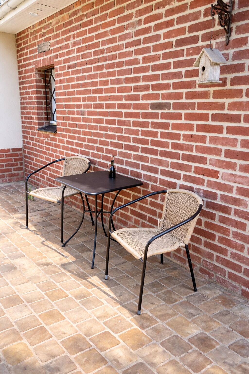 Um pátio com parede de tijolos, uma mesa, duas cadeiras de vime e um comedouro para pássaros.