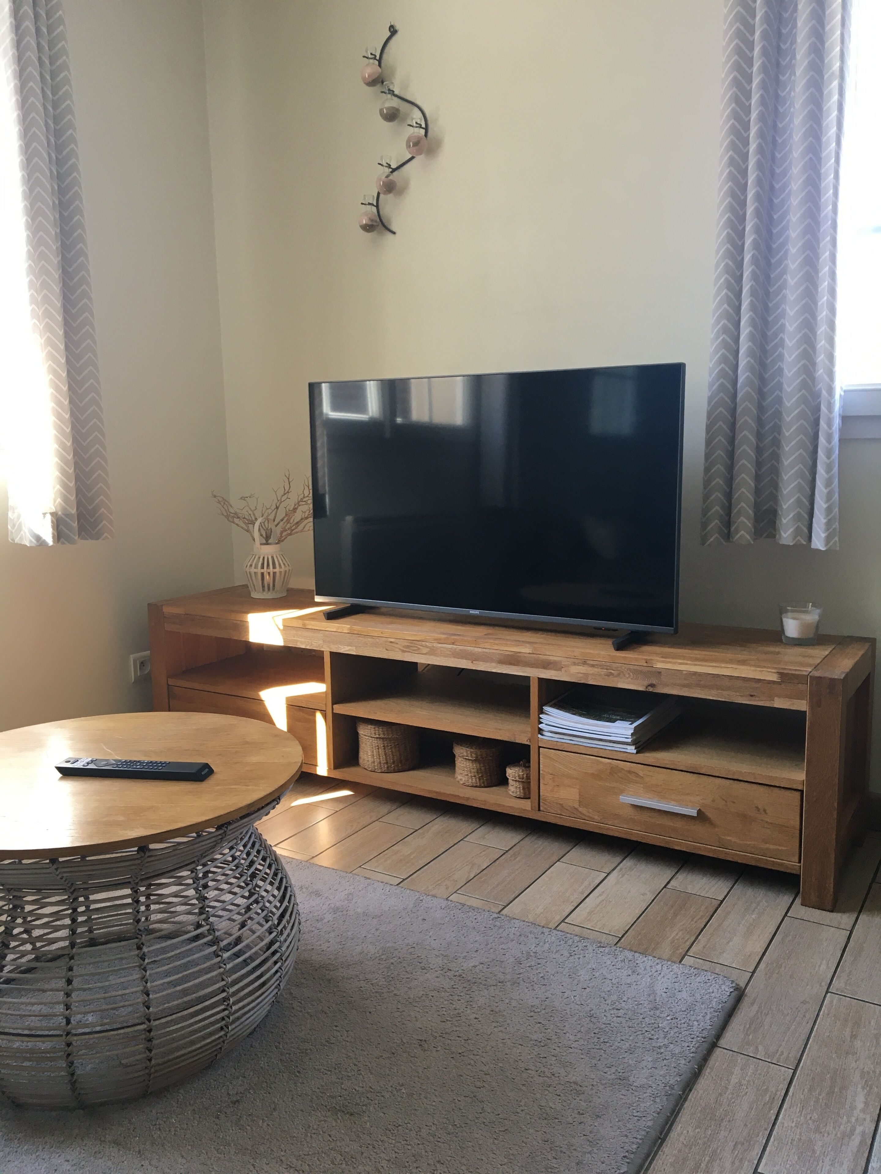 A living room with a flat-screen TV on a wooden stand, a round wooden table with a remote, a woven pouf, and a grey rug.
