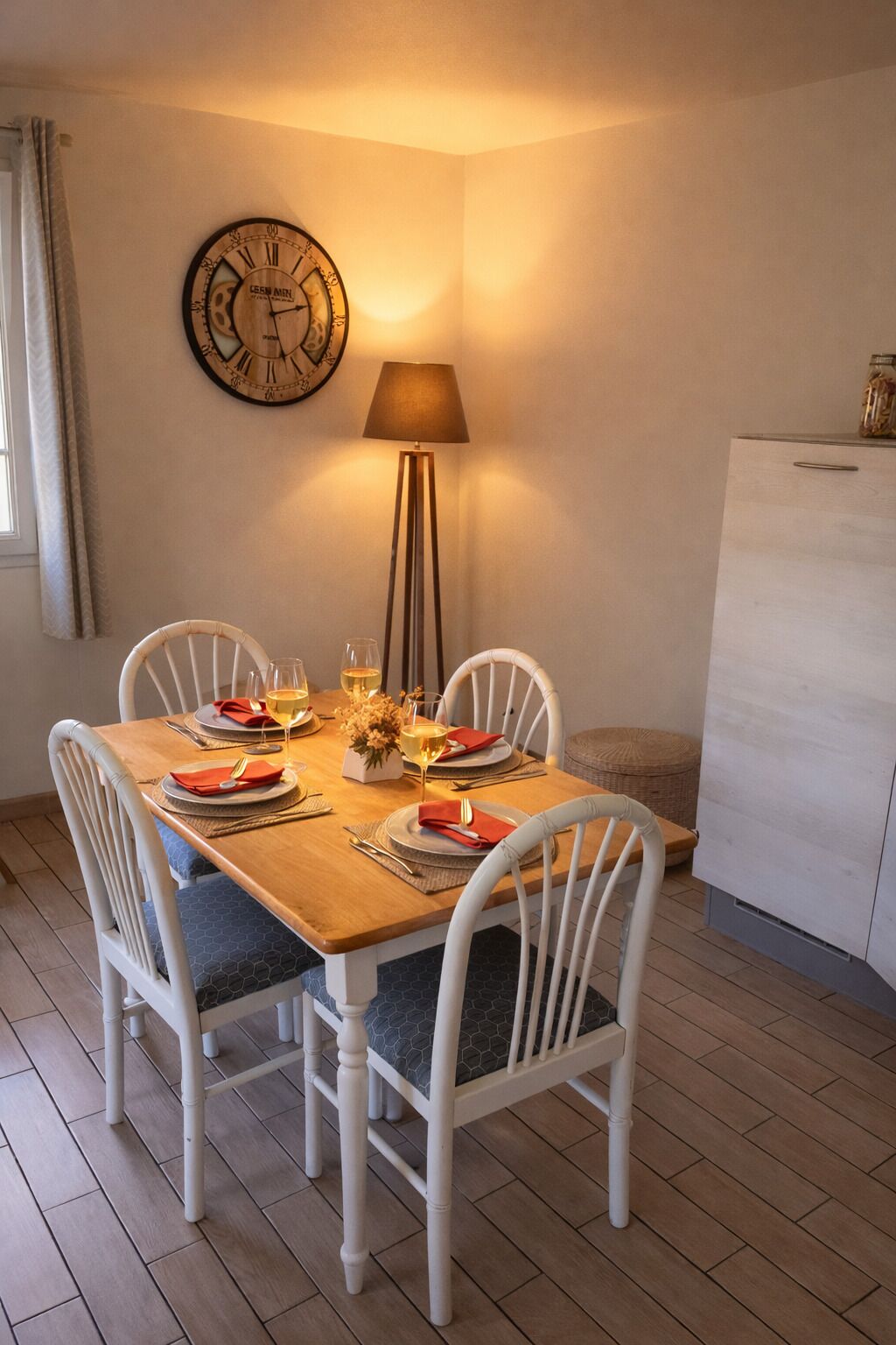 A dining area with a wooden table set for two, white chairs, a wall clock, a floor lamp, and a refrigerator.