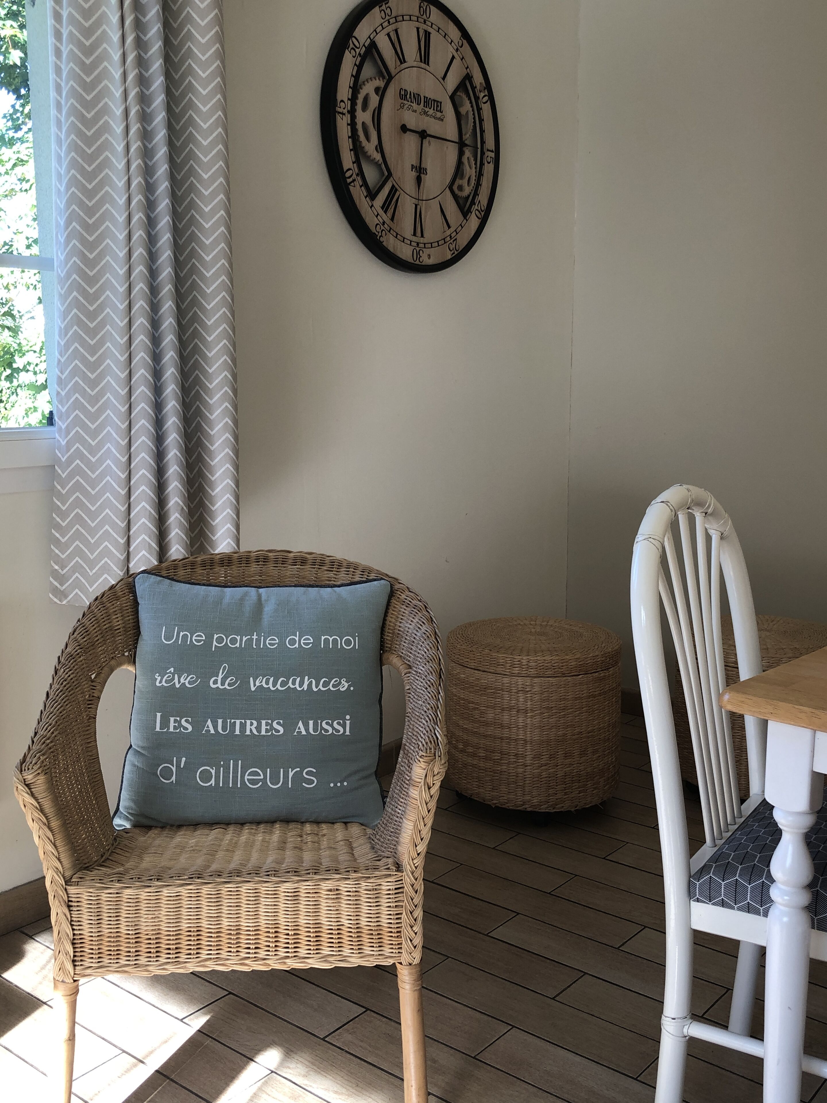 A wicker chair with a cushion, a white chair, a small round ottoman, and a wall clock with Roman numerals.
