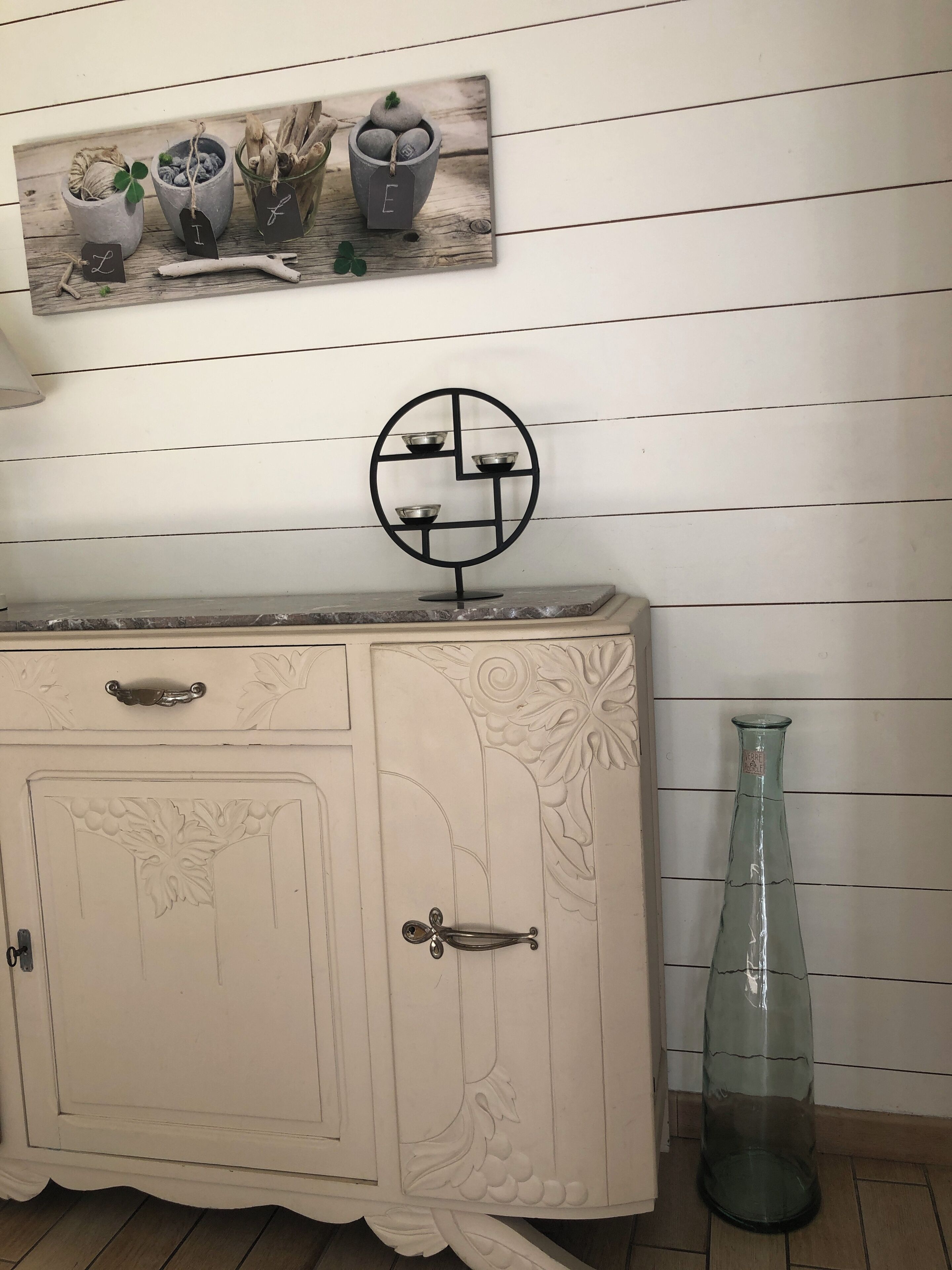 Un meuble blanc orné de motifs sculptés, une étagère en métal noir avec des bols, et une bouteille en verre transparent posée sur le sol.